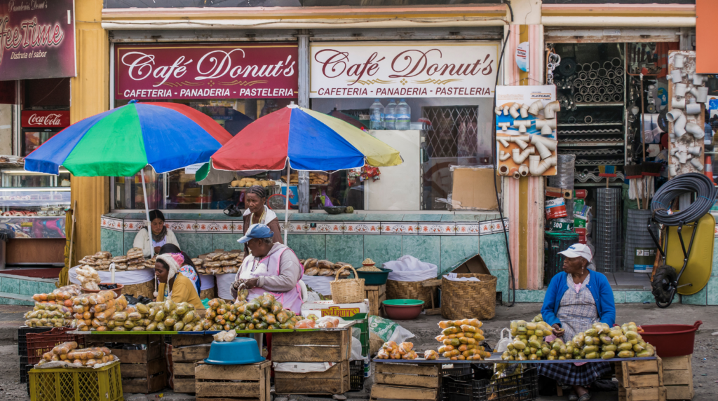 Illustration of a traveller at a bustling Latin American market stall, appearing slightly confused while pointing at fruit, with a friendly vendor nearby and signs suggesting different Spanish words for the same items, highlighting real-life language misunderstandings while travelling.